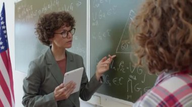 Kadın Matematik öğretmeni öğrencisinin önünde duruyor ve tahtaya yazılmış geometri problemini nasıl çözeceğini anlatıyor.