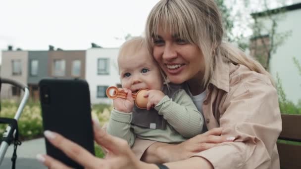 スマートフォンでビデオ通話で彼女のラップチャットで赤ちゃんと屋外に座っている陽気な若い白人女性の選択的な焦点ショット — ストック動画