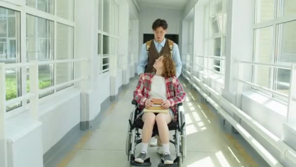 年轻的亚洲男子推着轮椅 让白人残疾女孩沿着大学走廊边走边聊天 — 图库视频影像
