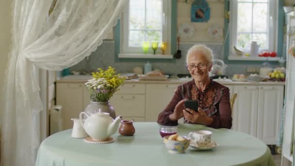 一位高个子白人妇女坐在乡村厨房的餐桌旁 享受着智能手机上的照片 — 图库视频影像
