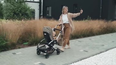 Bebek arabasında bebeğiyle yürürken blog için video çeken modern beyaz bir anne resmi.