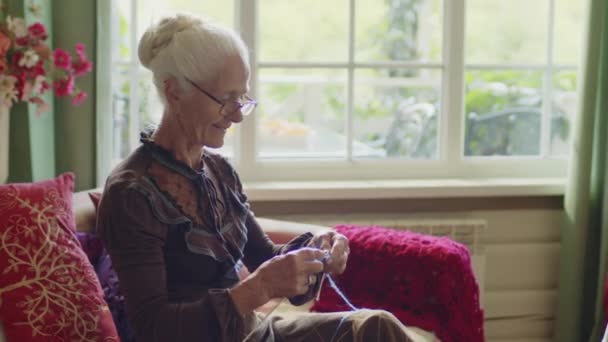 中等镜头的快乐奶奶穿着雅致的衣服和眼镜坐在沙发上编织什么东西 大妈美学 — 图库视频影像