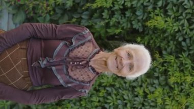 Kafkasyalı bir büyükannenin dikey portresi. Eski bir kıyafet giymiş. Duvara sarılmış, kameraya gülümseyen bir sarmaşıkla.