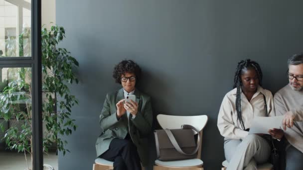 黒人女性とコーカサス人の男性が文書を議論しながら スマートフォンを使用して白人女性のミディアムフルショット すべてのインタビューを待っています — ストック動画