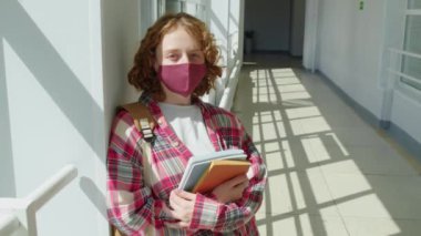 Okul koridorunda kameraya bakan koruyucu maske takmış, elinde kitaplarla tanınmayan kızıl saçlı bir kızın orta boy portresi.