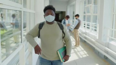 Kolej koridorunda durup kameraya bakarken koruyucu maske takan Afrikalı Amerikalı bir öğrencinin orta boy portresi.