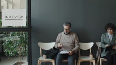 Yetişkin, beyaz gözlüklü, röportaj için bekleyen, CV 'sini tutan, kahve içen, diğer etnik çeşitliliğe sahip insanların yanına oturduğu, yanından ayrıldığı,