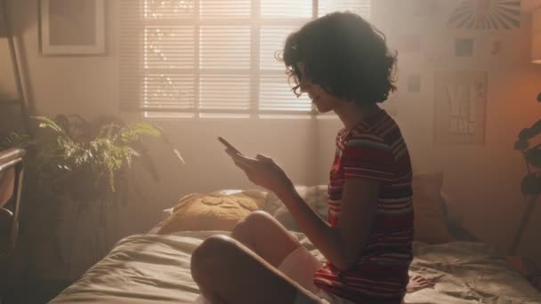 穿着亮晶晶T恤的短发少女坐在窗边未做的床上 用智能手机打字的侧影映入眼帘 — 图库视频影像
