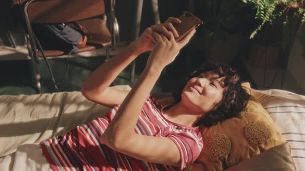 Kıvırcık Saçlı Çıplak Tişörtlü Genç Bir Kızın Yatakta Yatarken Telefonuna — Stok video