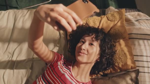 Kıvırcık Saçlı Parlak Tişörtlü Telefon Kamerasına Gülümseyen Yapılmamış Yatağında Uzanan — Stok video