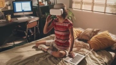 Beyaz VR kulaklıklı, parlak tişörtlü ve şortlu, elleri dizüstü bilgisayarda, başını oynatan kısa saçlı genç bir kız. Güneşli yatak odası