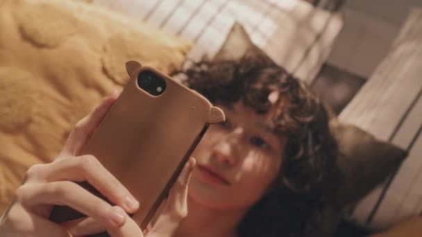 彼女のスマートフォンにタイピングカーリー短髪のティーンガールのハイアングルショット 朝の明るい寝室の枕に横たわる — ストック動画