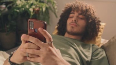 Yeşil tişörtlü, kıvırcık saçlı genç adamın sabahları yatağında yatarken telefonuna yazdığı gevşek yakın plan fotoğrafları. Arkaplanda yeşil bitki görünüyor
