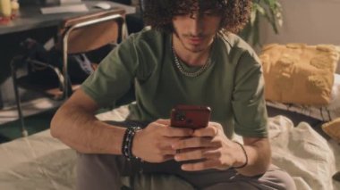 Orta boylu, kıvırcık saçlı, genç bir adamın gündüz vakti yatak odasında telefonuna yazı yazıp arka plandaki monitörü açtığı bir fotoğraf.