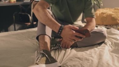 Koyu renk saçlı kıvırcık saçlı yeşil tişörtlü genç bir adamın sabahın köründe yapılmamış yatağında otururken aniden telefonunu kontrol ederken sonra da kenara fırlatırken. Güneşli yatak odası, ekran arkası