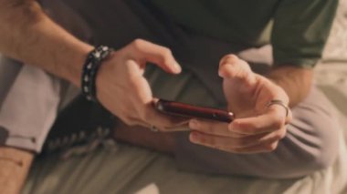 Yeşil tişörtlü genç adamın orta bölüm gayri resmi. Yatakta oturuyor, akıllı telefonuna tıklıyor. Ön planda erkek mücevherleri var.