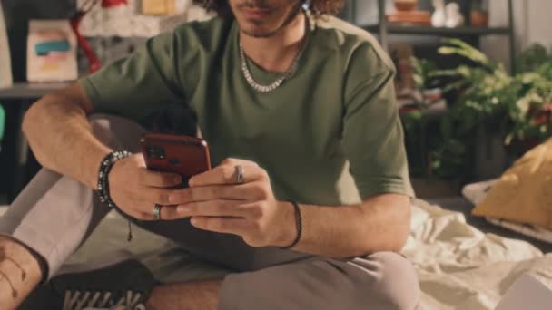 一个身穿绿色T恤 头戴男性珠宝的年轻人坐在未做完的床上 在智能手机上点击了一下 背景为阳光明亮的卧室 绿色植物及书桌 — 图库视频影像