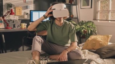 Beyaz VR kulaklıklı kıvırcık bir adamın orta boy fotoğrafı. Başını yavaşça çeviriyor. Önündeki dizüstü bilgisayarla dağınık bir yatakta oturuyor. Ekranı aç, arka planda lambalar ve pencere, sabahın erken saatlerinde.