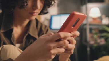 Kahverengi ceketli kıvırcık kızın telefonunu kontrol edip kaldırırken, üzgün görünürken, yatakta otururken, ekranı ve arka plandaki bulanık lambayı açarken.