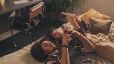 Genç çiftin telefonlarıyla yatakta yatıp ekranlarını birbirlerine gösterip gülümsedikleri yüksek açılı bir çekim. Arka planda bitki ve sandalye olan bir masa