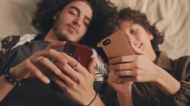 Genç erkek arkadaş ve kız arkadaş telefonlarıyla yatakta uzanıp birbirlerine bakıyorlar, konuşuyorlar, gülüyorlar. Güneş ışığı, üst görünüm