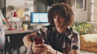 Telefonuyla yazı yazan kıvırcık bir gencin orta boy portresi. Sonra odasında yatağında otururken kameraya ciddi ciddi bakıyor. Arka planda ekran, bitki ve lamba açık.