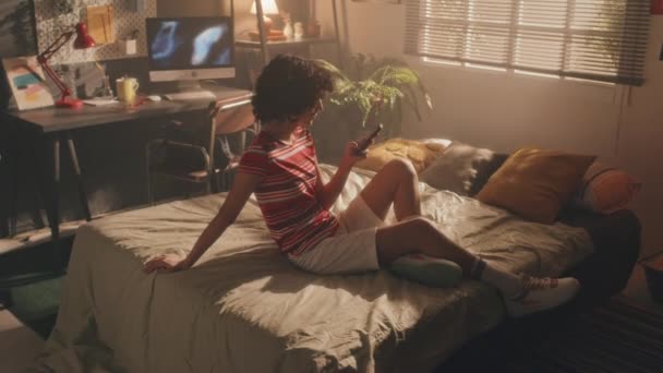 满满一帧卷曲的少女坐在房间中央的阳光下的床上 在电话上打字 桌子上有打开的监视器屏幕 背景中的窗户 — 图库视频影像