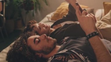 Genç bir adam ve genç bir kız, ikisi de kıvırcık, telefonlarıyla yatakta uzanıyor, birbirlerine bakıyor, gülümsüyorlar. Yüksek açı görünümü