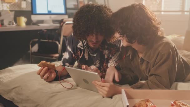 中等比例的卷曲少女和小伙子躺在床上 头戴平板电脑 戴着耳机 吃着披萨 享受着自己的生活 背景中打开监视器和窗户 卧室里阳光灿烂 — 图库视频影像