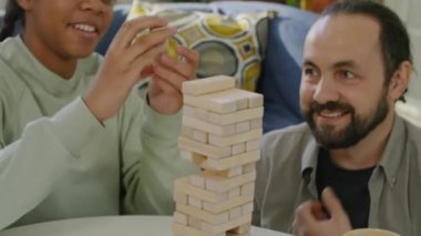 Siyah kız ve beyaz adamın aydınlık odada masa oyunu oynadığı orta boy bir fotoğraf. Kız blok ekliyor, adam dikkatlice alıyor, kule düşüyor, kız ve erkek gülüyor, arkalarında mavi kanepe var.