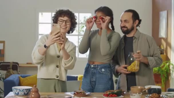 满满的中镜头是白种人的眼镜年轻女子 黑人少女和白种人男子 他们站在餐桌前 端着盘子和食物 三个女孩都在为自己笑 一个女孩把樱桃戴在眼睛上 — 图库视频影像