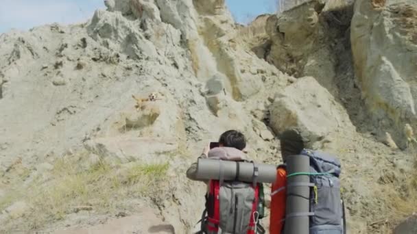 两名女游客在岩石脚下站着背包 其中一人拍摄了他们的狗在略高一点的地方跑来跑去的视频或照片 刮风的天气 后视镜 — 图库视频影像
