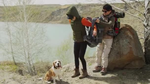 全景拍摄到两名亚洲朋友站在岩石湖畔的岩石边 其中一人帮助另一人把背包放在旁边 一只小狗坐在旁边 风和日丽 — 图库视频影像