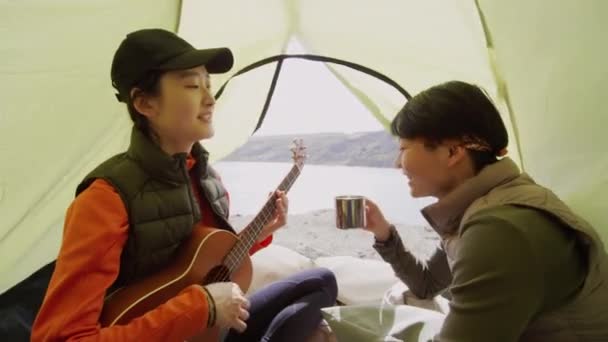 Две Азиатские Туристки Сидят Палатке Берегу Озера Одна Играет Гитаре — стоковое видео