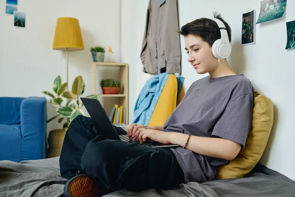 ラップトップスクリーンを見て ベッドに座って学校のプロジェクトで働いている間 野心的な音楽を聴くヘッドフォンの十代の少女 — ストック写真