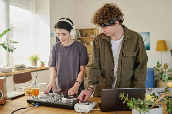 現代の十代のカップルは新しいコンピュータ音楽を作成し 家の環境でターンテーブルとラップトップが付いている机によって立っている間それを録音します — ストック写真