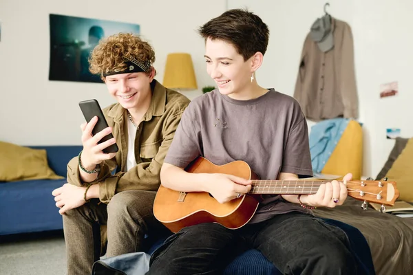 Cheerful teenage couple in casualwear looking at smartphone screen and taking selfie or shooting video while girl playing acoustic guitar