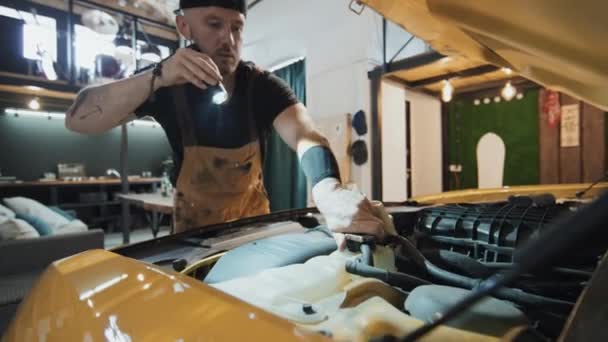 汽车机械师在车盖中打开油箱的中镜头 用火把点燃 车库内部 人工照明 沙发和桌子背景 — 图库视频影像