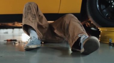 Sarı araba gövdesinin altında yatan mekanik bacakları kapatın. Onarımlar yapıyor. Yanında birkaç alet var.