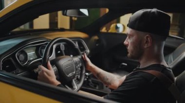 Sarı araba motorunu çalıştıran, direksiyonu tutan, mutlu bir gülümsemeyle geriye yaslanan, garajın içindeki dövmeli adamın yan tarafları gevşemiş.