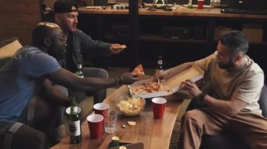 Üç çok ırklı arkadaşın oturma odasında takılıp, kutudan pizza dilimleri alırken, gülüşürken, konuşurken, DJ 'lik yaparken, oturma odasında bira içerken tam orta ölçekli bir fotoğraf.