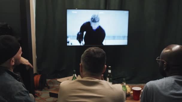 三个多种族的男性朋友坐在客厅的沙发上 坐在大屏幕电视上观看冰球比赛 他们背对着面前的啤酒瓶和点心 情绪反应热烈 — 图库视频影像