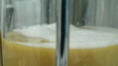 Bira demleme işlemi sırasında cam kaba doldurulan bulutlu sarımsı bira otunun yakın çekim görüntüsü, üzerinde yağ ve köpük var, örnekleme ve kalite kontrolü için.