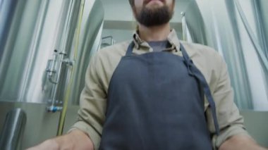 Beyaz sakallı, önlüklü, bira fıçısı taşıyan, üretim katında paslanmaz çelik fermantasyon tankları taşıyan beyaz erkek işçinin orta boy yakın çekimi.