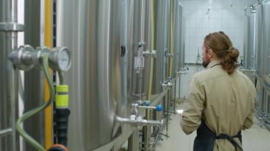 Bira fabrikasında bira mayalama tankları, apronlu beyaz sakallı erkek teknisyen içeri giriyor, basınç göstergesine basıyor, tablet üzerine notlar alıyor ve bir sonrakine geçiyor.