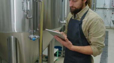 Geniş brite bira tankının yanında duran, basınç göstergesine bakan ve tablet bilgisayardaki kontrol listesine not alan beyaz erkek bira üreticisinin orta boy görüntüsü.