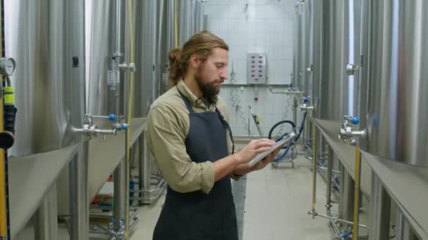 中弧线拍摄的年轻红头发白种人男性酿酒厂工人 留着胡子 站在啤酒发酵罐附近 看仪器 并在平板电脑上填写检查清单 — 图库视频影像