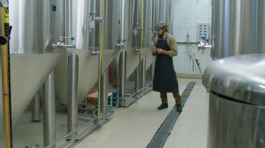 Bira fabrikasında üretim tesisi boyunca önlüklü ve kapşonlu genç beyaz erkek teknolojinin tam karesi, parlak bira tanklarındaki ölçümleri kontrol etmek ve tablet bilgisayar raporunu tamamlamak.