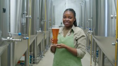 Afrika kökenli Amerikalı kadın bira imalathanesi teknisyeninin çelik tanklar arasında fermantasyon odasında dururken çekilmiş orta boy portresi. Elinde yeni demlenmiş bira bardağı ve kameraya gülümsüyor.