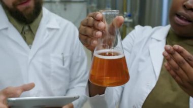 Yakın plan cam matara, yeni üretilen bira örnekleri ve arka planda beyaz önlüklü iki anonim çoklu etnik kökenli bira fabrikası çalışanı, doku, tat ve lezzeti tartışıyorlar.